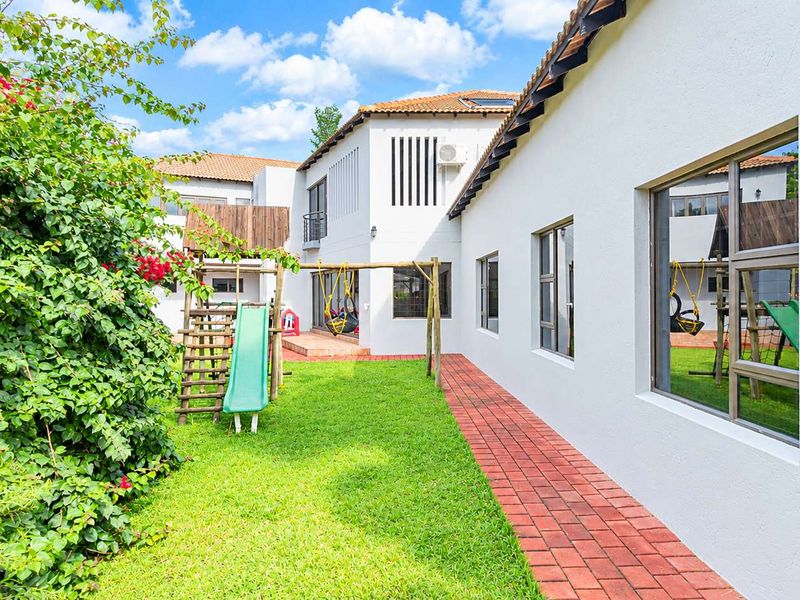 A well-maintained rear exterior view of the house featuring a brick paved walkway, lush lawn, and clean white facade with aluminium window frames.