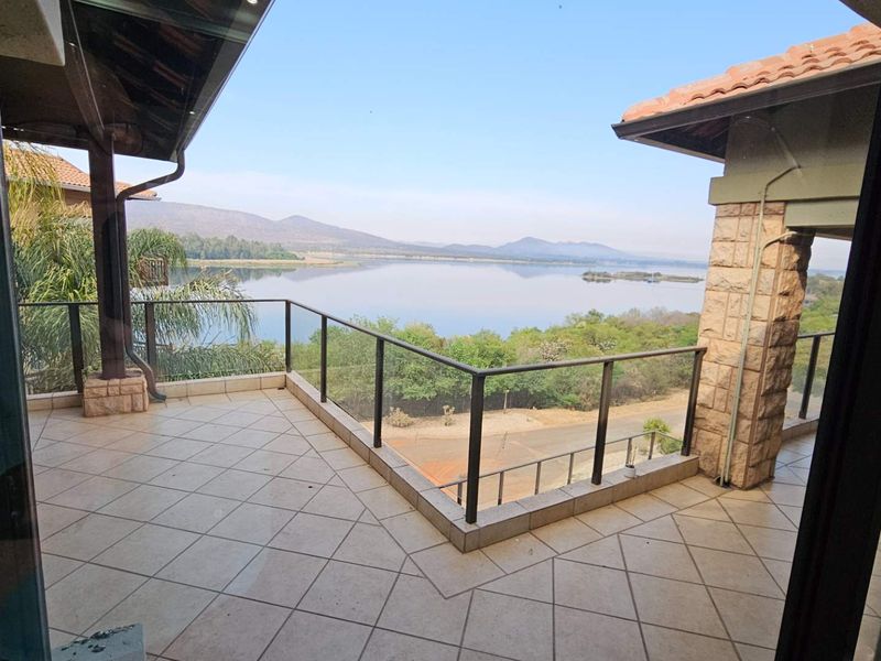 Spacious covered balcony featuring tiled flooring, stone-clad pillars, and a glass balustrade, offering expansive views of the lake and surrounding landscape.