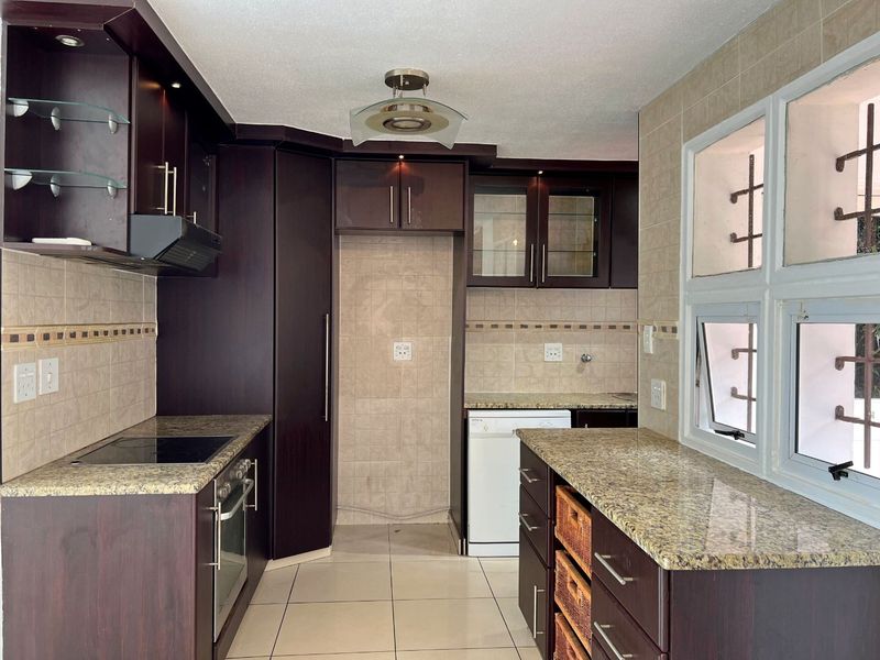 A functional kitchen featuring dark wood cabinetry, granite countertops, built-in oven, electric hob, and tiled finishes with security bars on the windows.