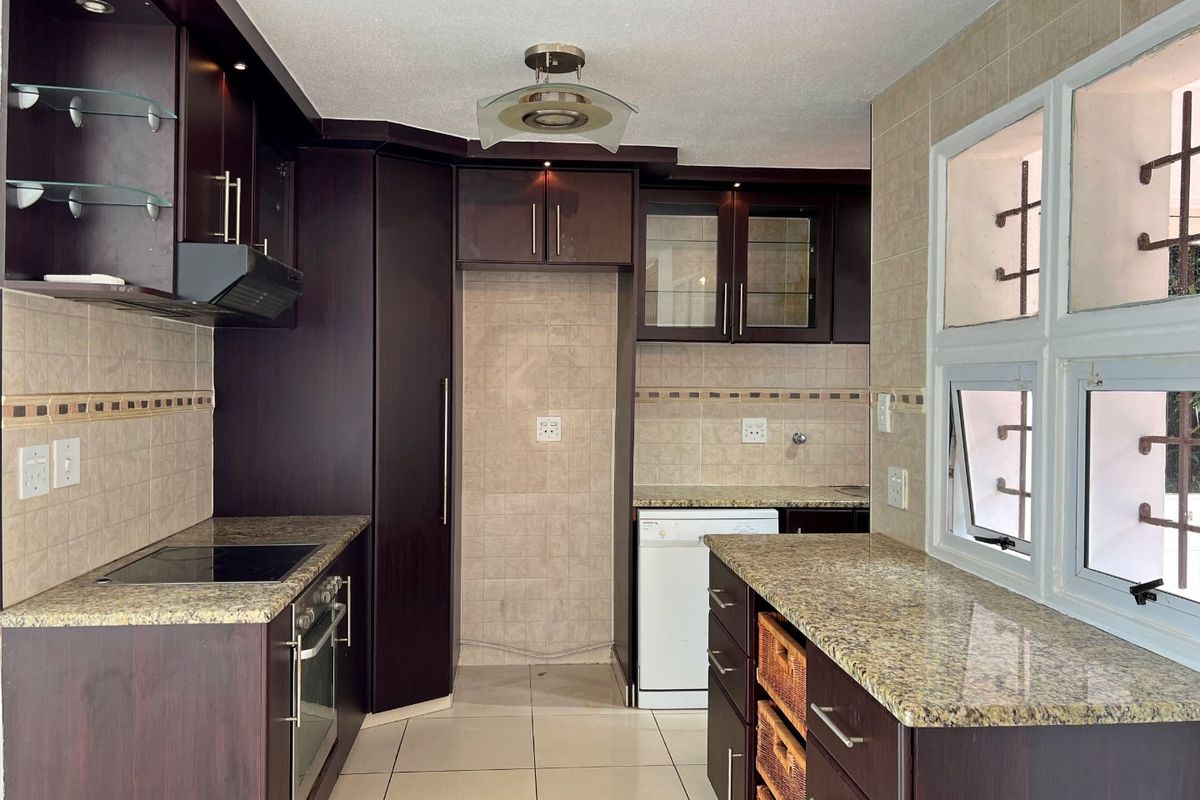 A functional kitchen featuring dark wood cabinetry, granite countertops, built-in oven, electric hob, and tiled finishes with security bars on the windows.