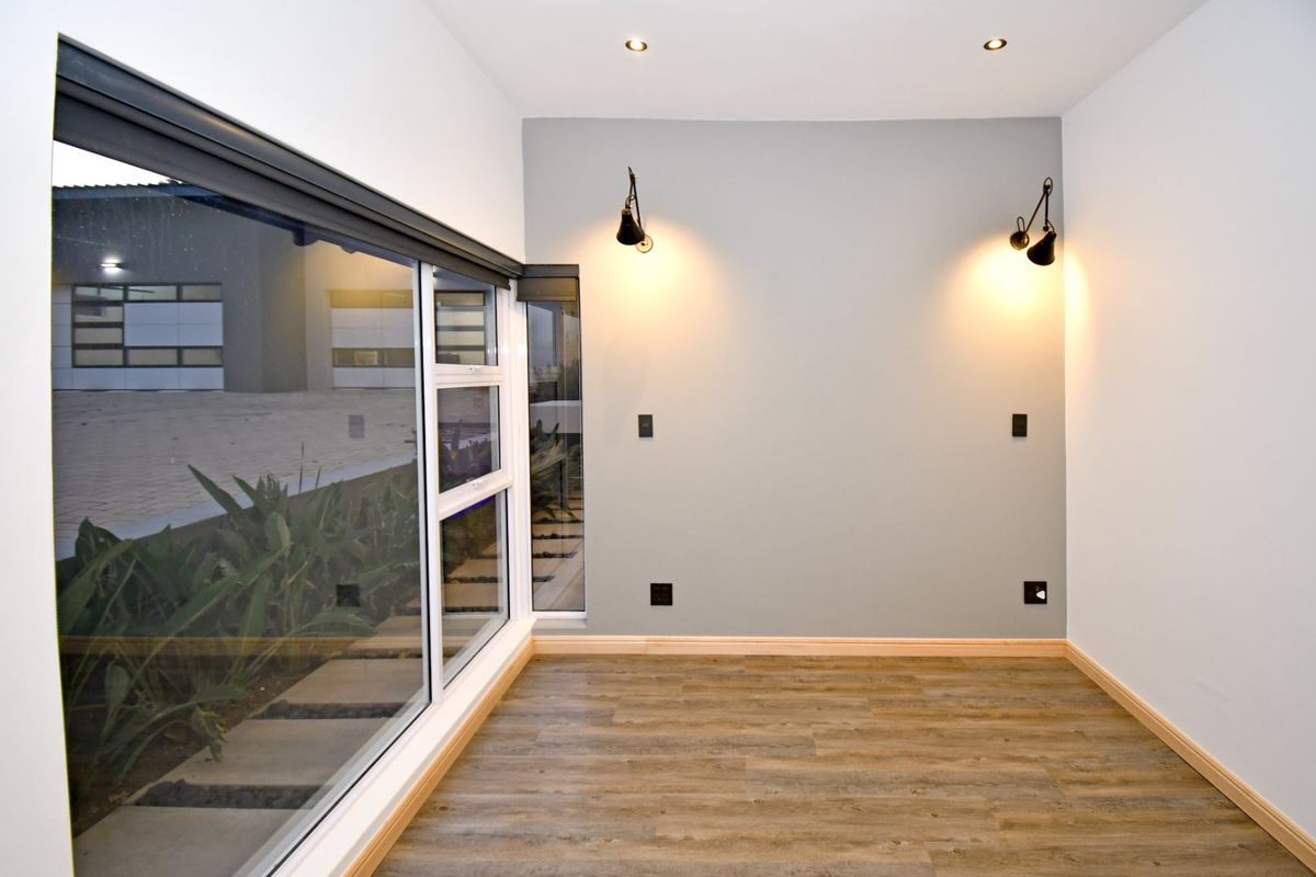 A bright, empty bedroom featuring modern laminate flooring, recessed downlights, wall-mounted accent lighting, and a large corner window overlooking the exterior.