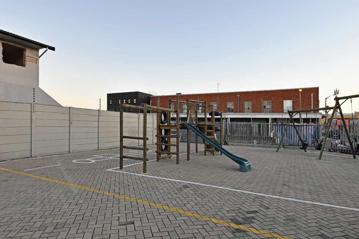 An outdoor paved courtyard area featuring a precast boundary wall with electric fencing and fixed playground equipment, adjacent to a building with unfinished exterior wall sections.