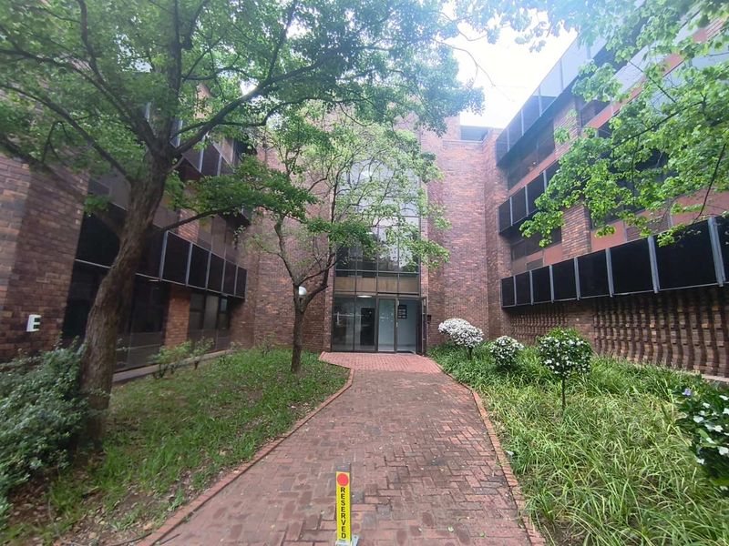 Front entrance view of a multi-story office building featuring a brick facade, glass entrance, and a paved walkway through a landscaped garden.