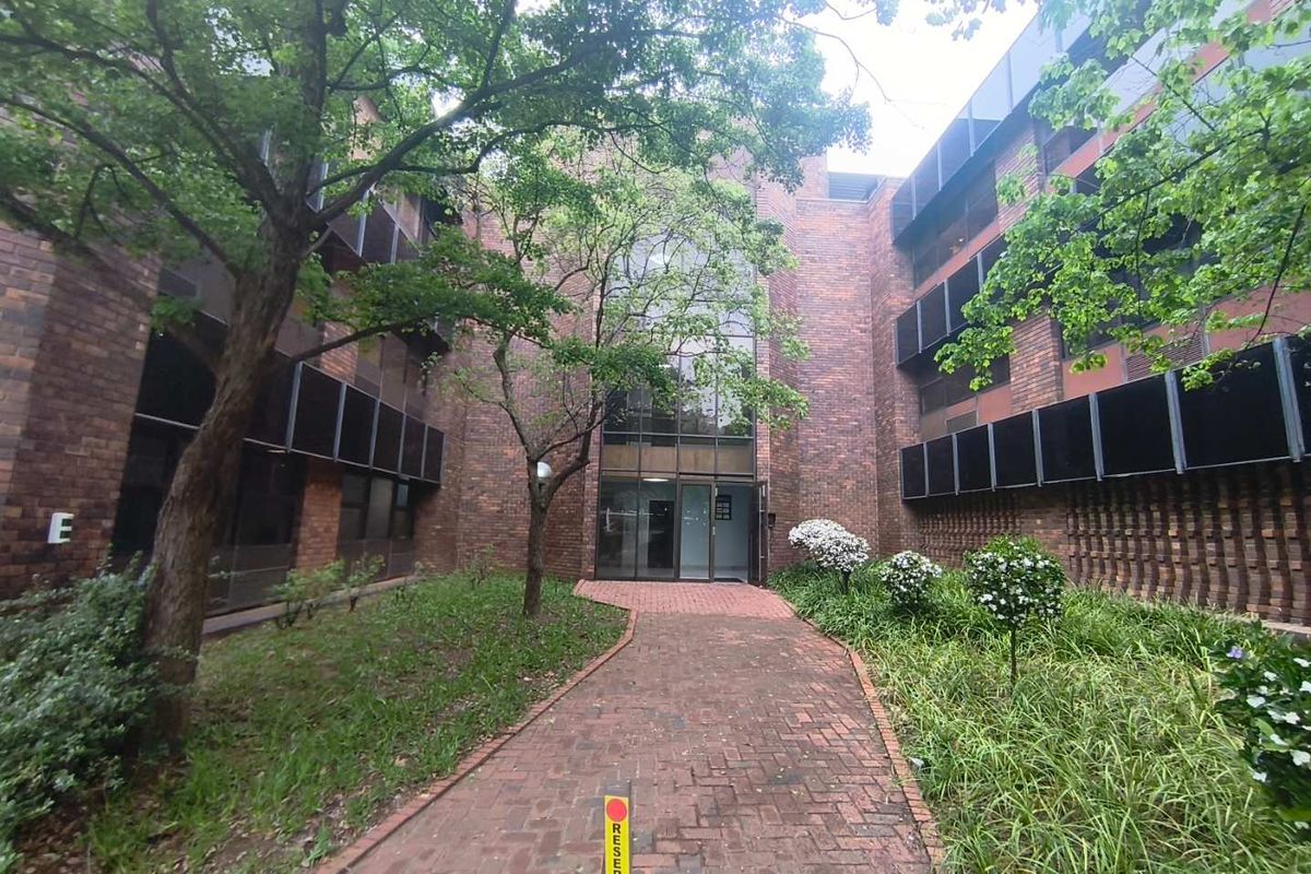 Front entrance view of a multi-story office building featuring a brick facade, glass entrance, and a paved walkway through a landscaped garden.