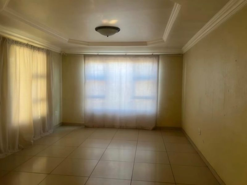 A spacious, empty living room featuring tiled flooring, a tray ceiling with decorative molding, and natural light through large windows.
