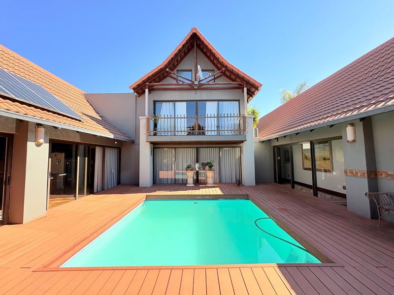 A well-maintained outdoor pool area featuring extensive timber decking, a central swimming pool, and a two-story building structure with a balcony.
