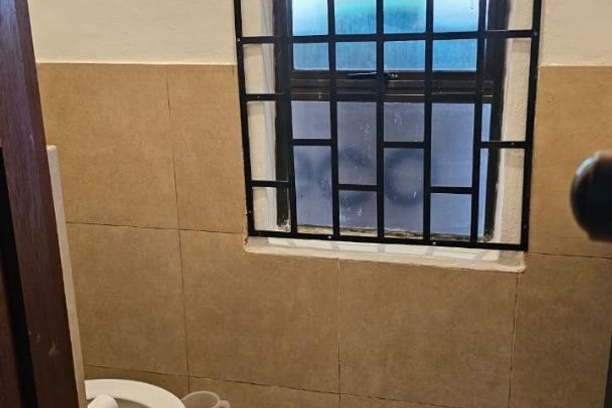 A small bathroom featuring tiled walls and flooring, equipped with a toilet and a window secured with metal burglar bars.