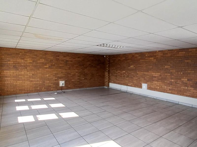 An open-plan industrial office space featuring exposed brick walls, tiled flooring, a suspended ceiling, and a wall-mounted air conditioning unit.