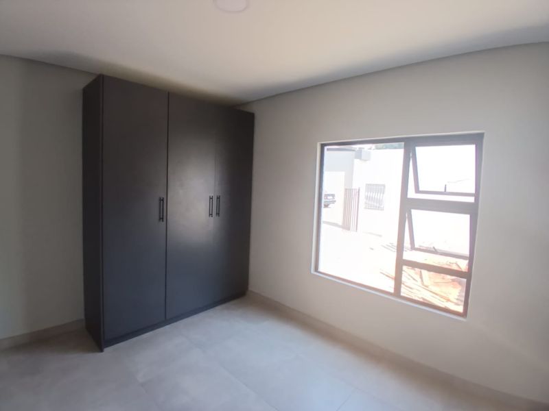 A bright, modern bedroom featuring neutral tiled flooring, a dark built-in wardrobe, and an aluminium window frame.