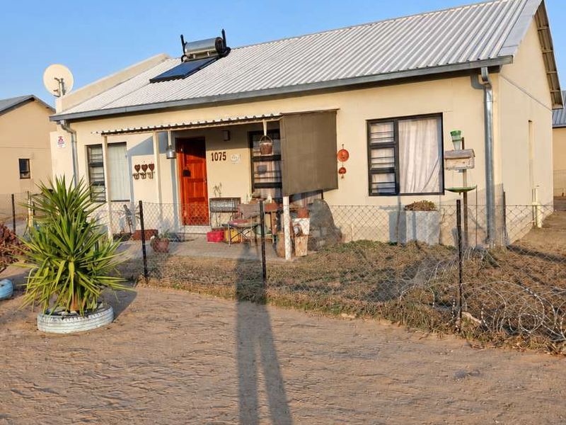 Front elevation of a single-story house featuring a corrugated metal roof, solar water heater, and perimeter fencing.
