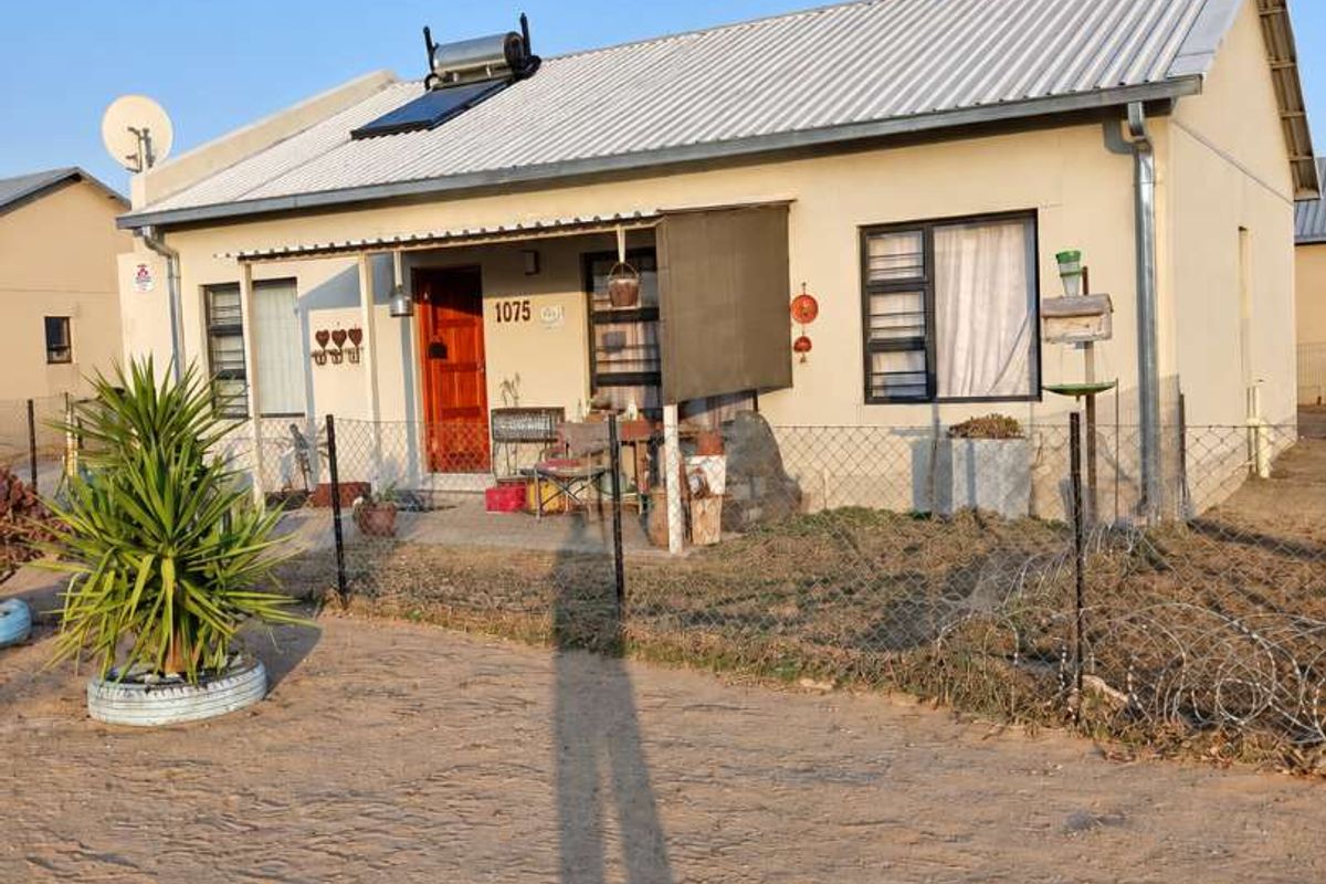 Front elevation of a single-story house featuring a corrugated metal roof, solar water heater, and perimeter fencing.