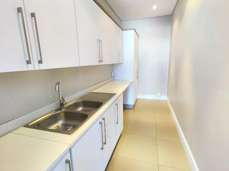 A clean, modern galley-style kitchen featuring white cabinetry, stainless steel double sink, and neutral tiled flooring.