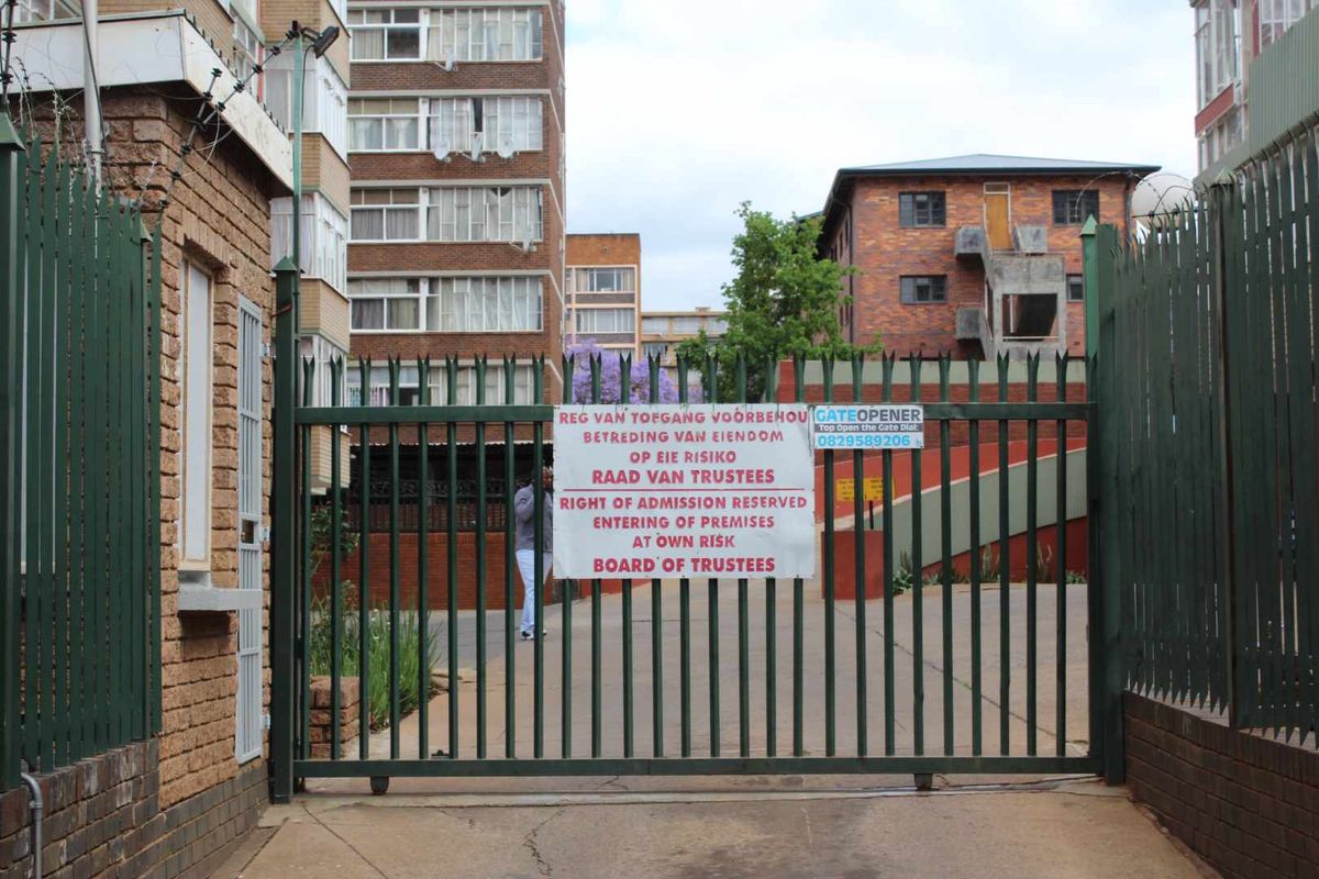 View of the apartment complex entrance featuring a secure metal gate, electric fencing, and paved driveway access.