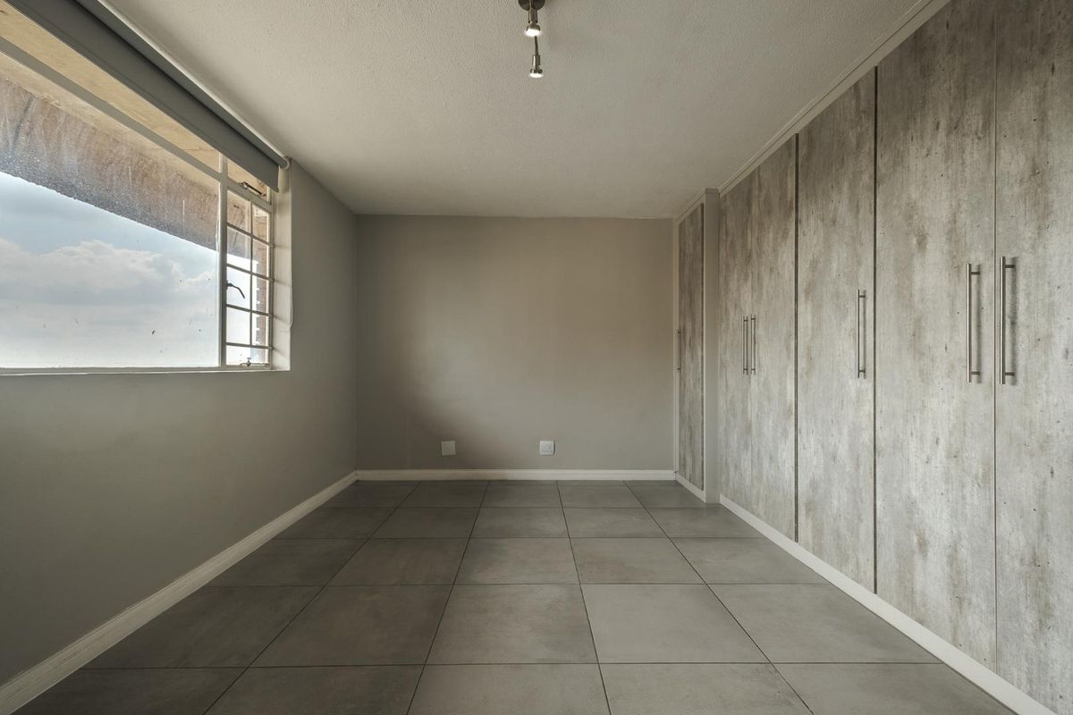 A bright, empty bedroom featuring modern grey floor tiles, neutral walls, and extensive built-in cabinetry with a contemporary textured finish.