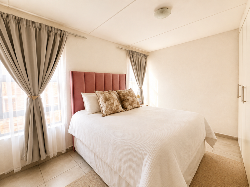 A bright and well-maintained bedroom featuring tiled flooring, built-in wardrobes, and large windows with neutral curtains.