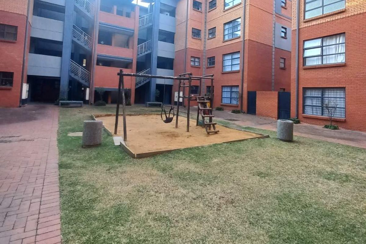 A communal courtyard area within an apartment complex featuring a brick facade, paved walkways, and a small playground set on a grassed area.