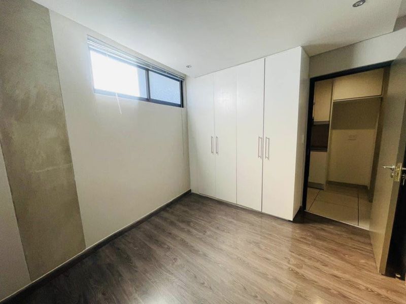 A clean, empty bedroom featuring modern laminate flooring, built-in white cupboards, and a high-level window providing natural light.