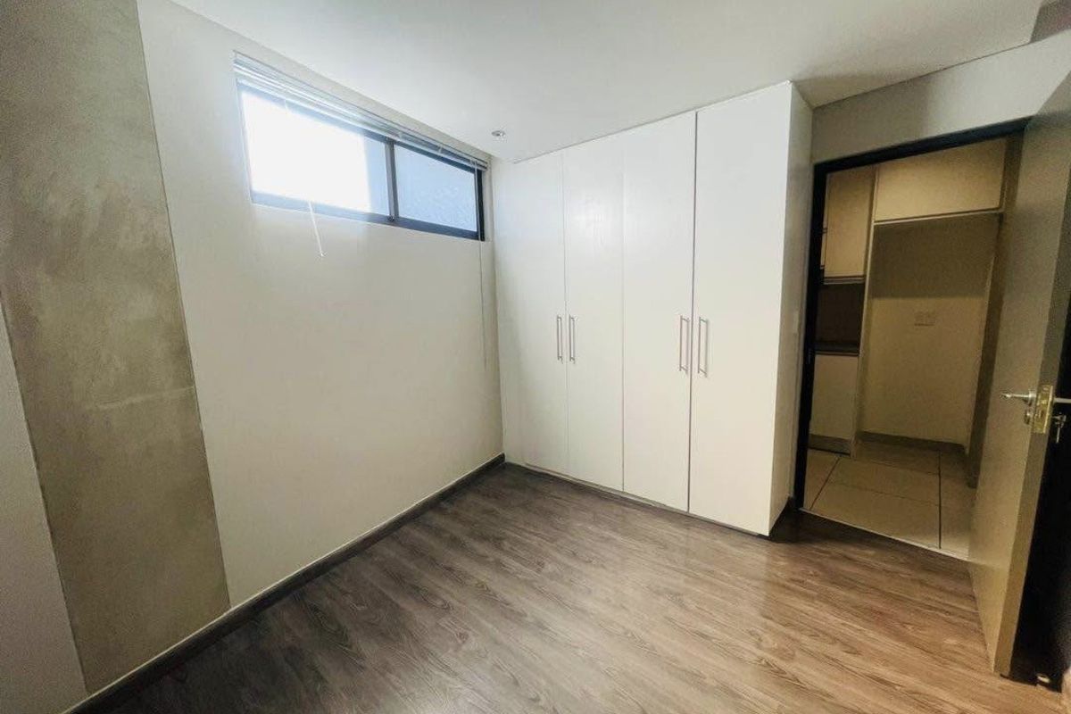 A clean, empty bedroom featuring modern laminate flooring, built-in white cupboards, and a high-level window providing natural light.