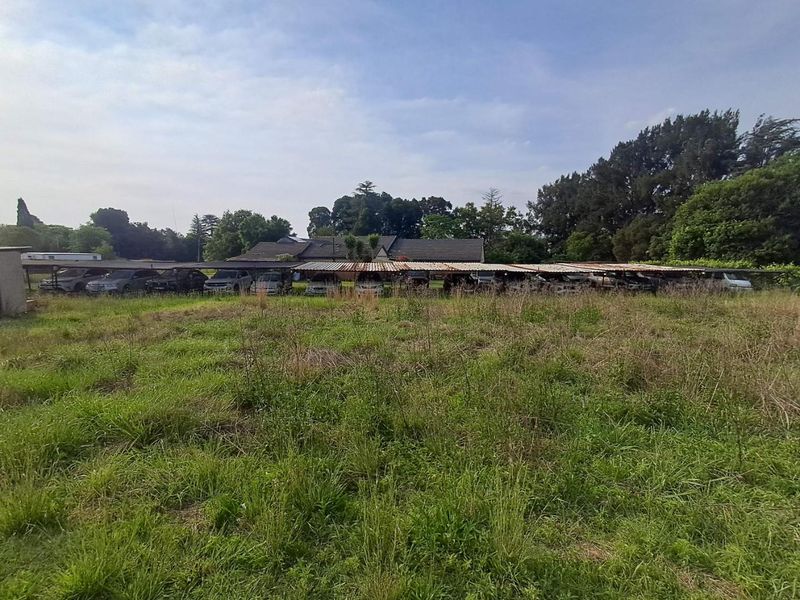 View of a large open field leading to a long, weathered carport structure with corrugated metal roofing on the property grounds.