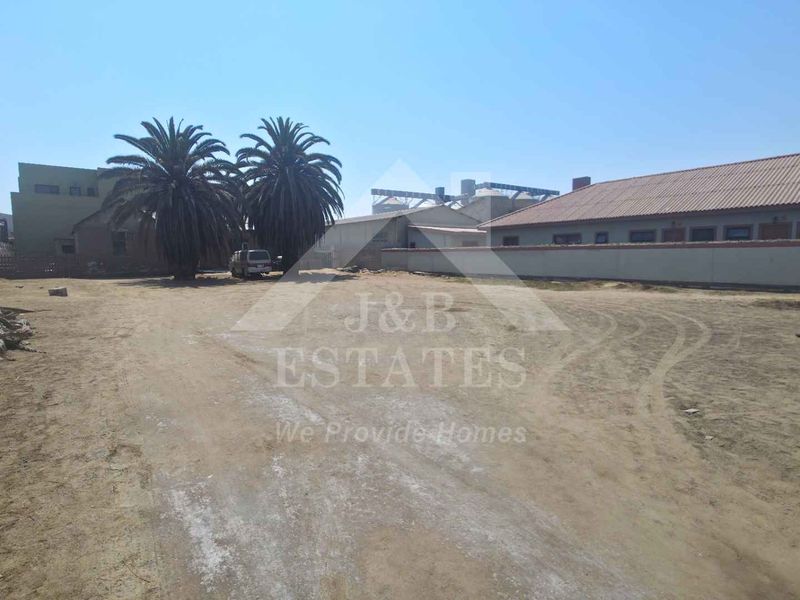 A large, unpaved industrial yard area featuring a perimeter wall and views of adjacent industrial structures.