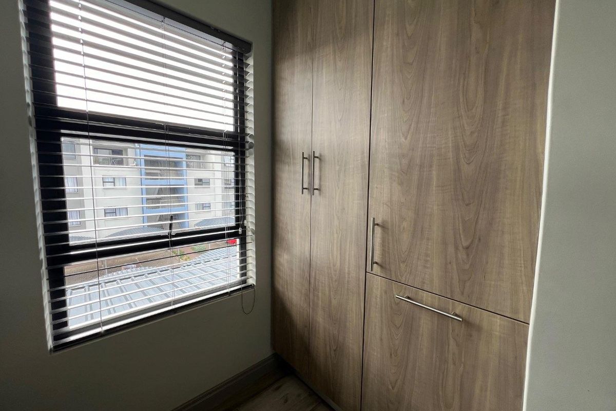 A clean bedroom featuring modern wood-finish built-in cupboards, laminate flooring, and a window fitted with venetian blinds.