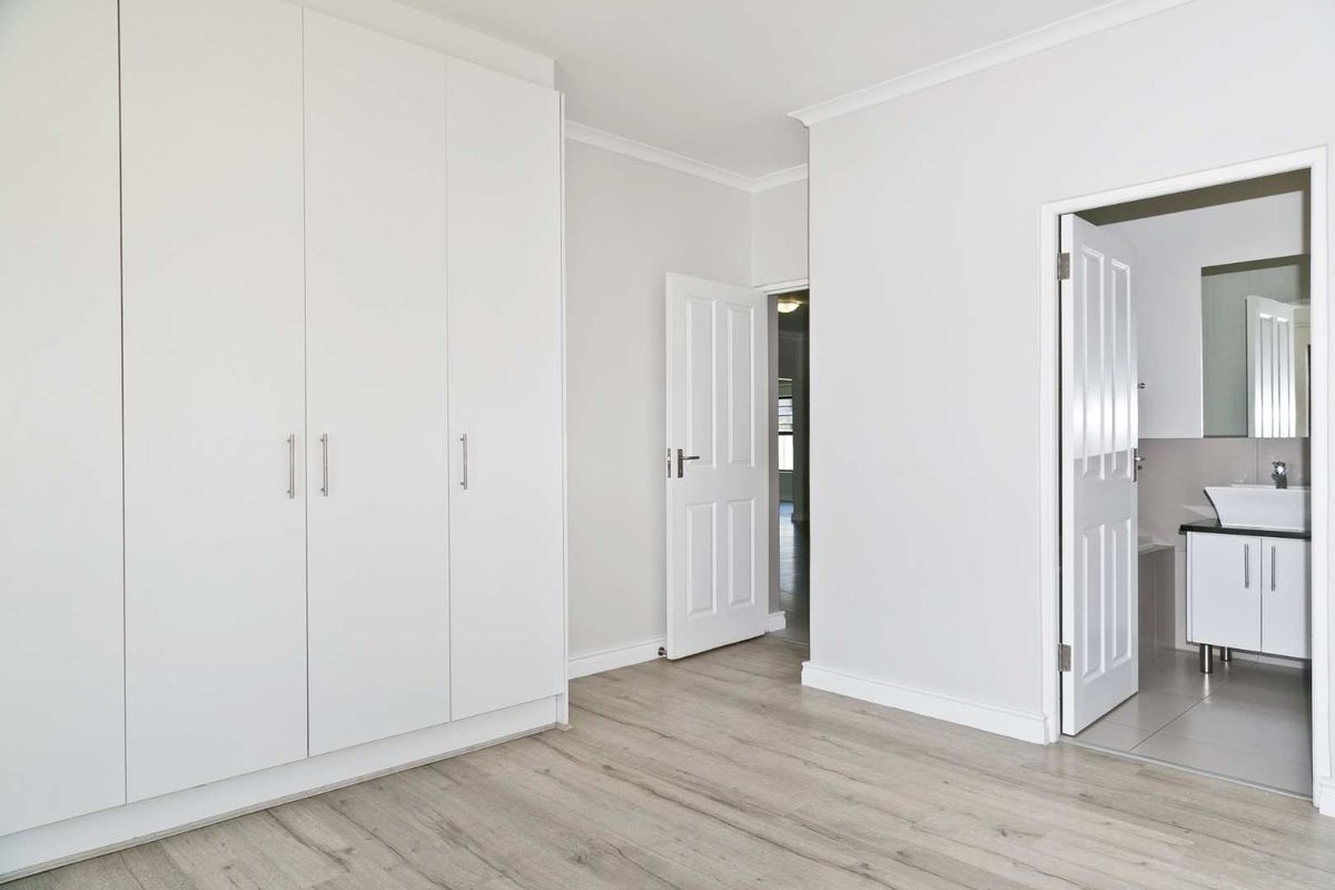 A bright, empty bedroom featuring modern laminate flooring, floor-to-ceiling built-in cupboards, and direct access to an en-suite bathroom.