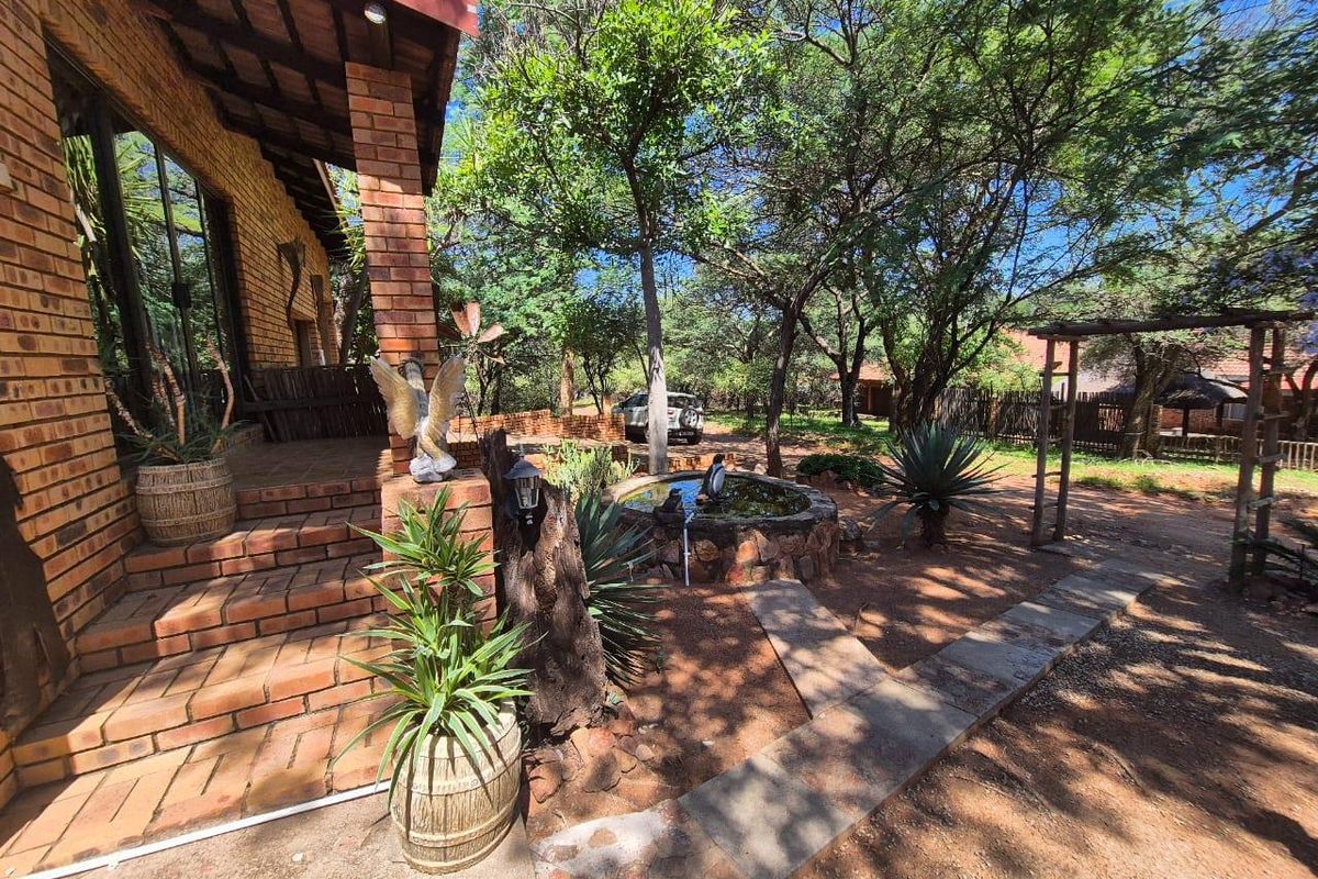 A rustic brick-built home featuring a covered patio, stone-clad water feature, and established garden with mature trees and natural pathways.