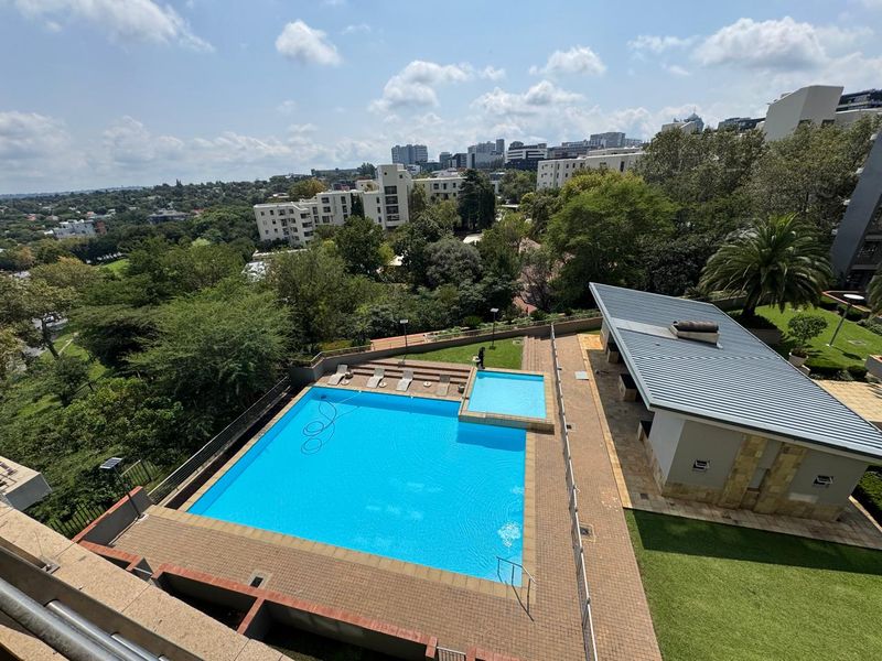 Elevated view of a well-maintained communal swimming pool area featuring a paved deck, adjacent clubhouse structure, and landscaped garden surroundings.