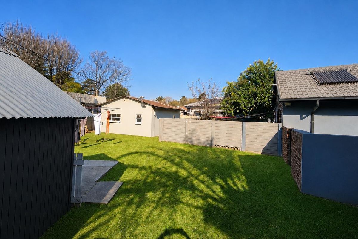 A view of the rear garden area featuring a lawn, concrete patio, and boundary walls, with a secondary structure and main house roof visible.