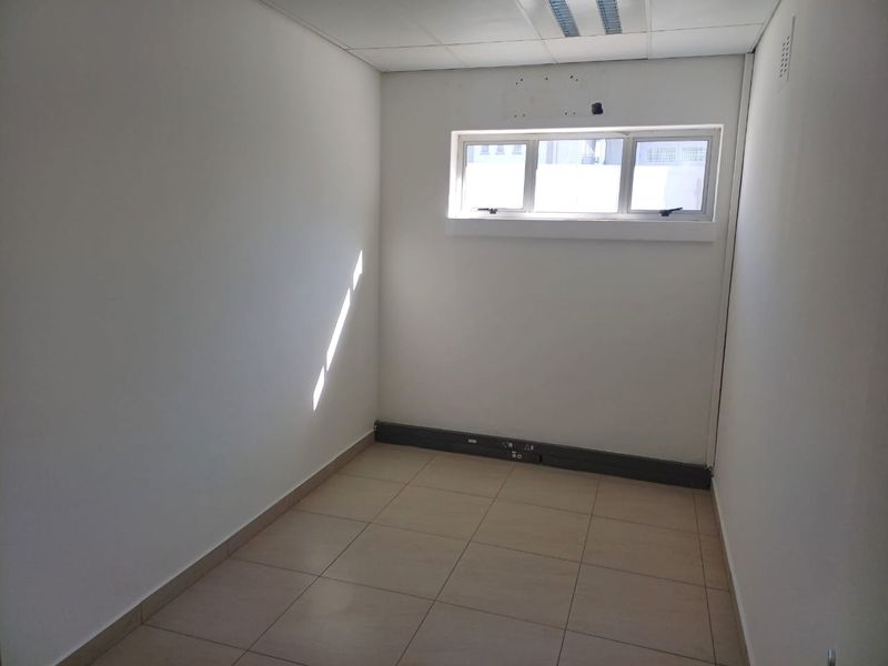 A vacant, compact office space featuring tiled flooring, a suspended ceiling with recessed lighting, and a horizontal window.