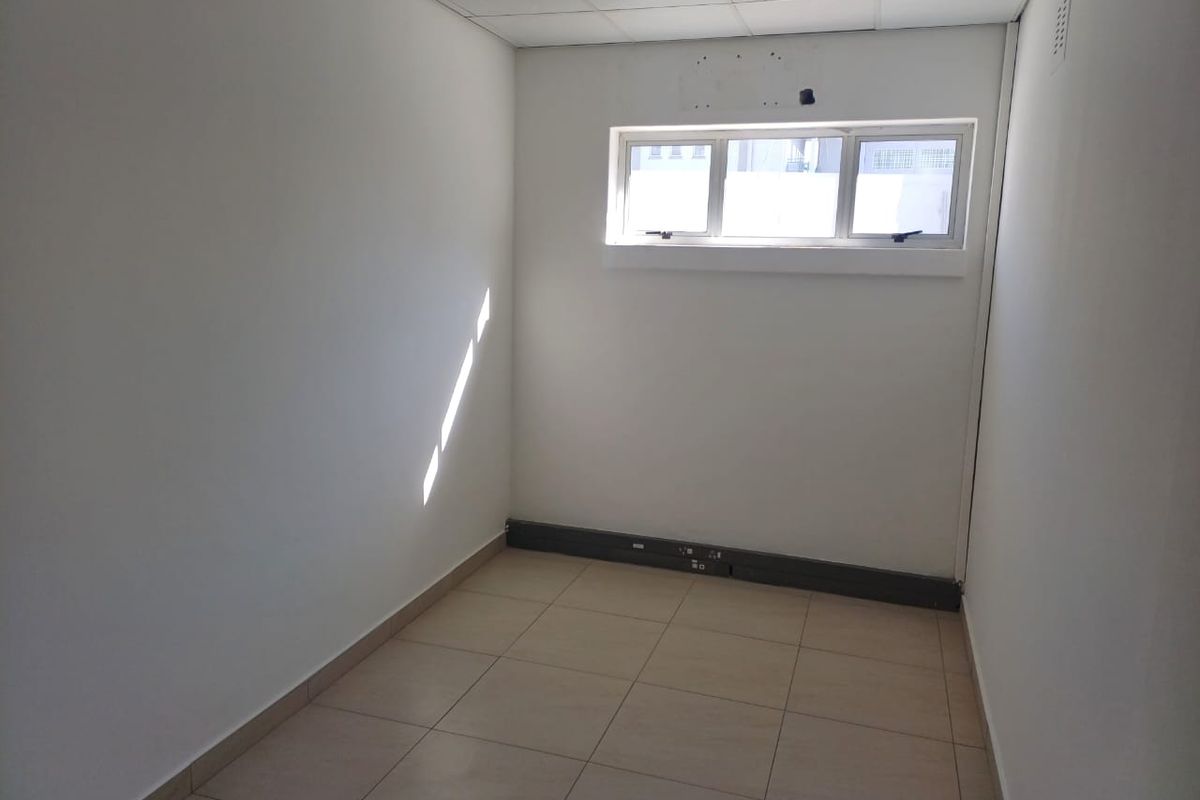 A vacant, compact office space featuring tiled flooring, a suspended ceiling with recessed lighting, and a horizontal window.