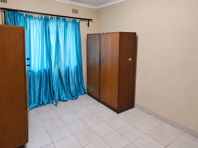 A functional bedroom featuring neutral tiled flooring, plain walls, and a curtain rod, offering a blank canvas for personalization.