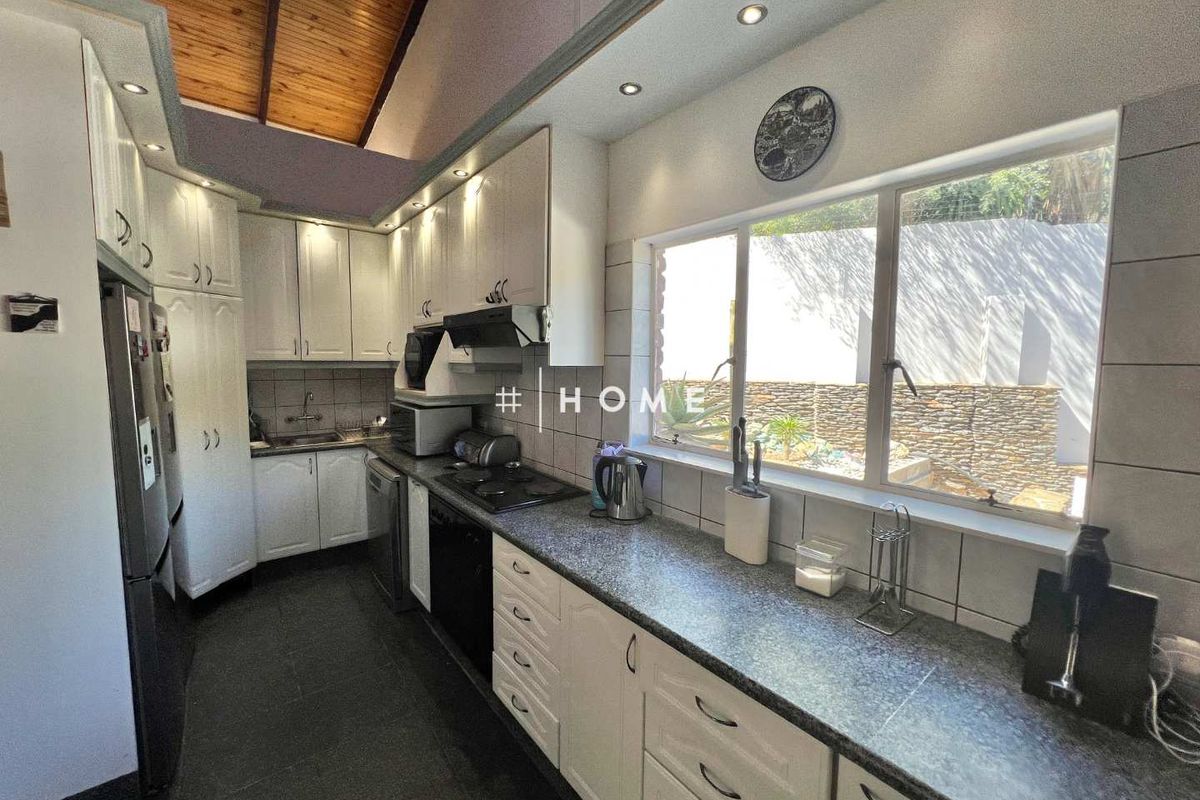 A functional kitchen featuring white cabinetry, granite countertops, a built-in hob, and a vaulted timber ceiling, overlooking a private rear garden area.