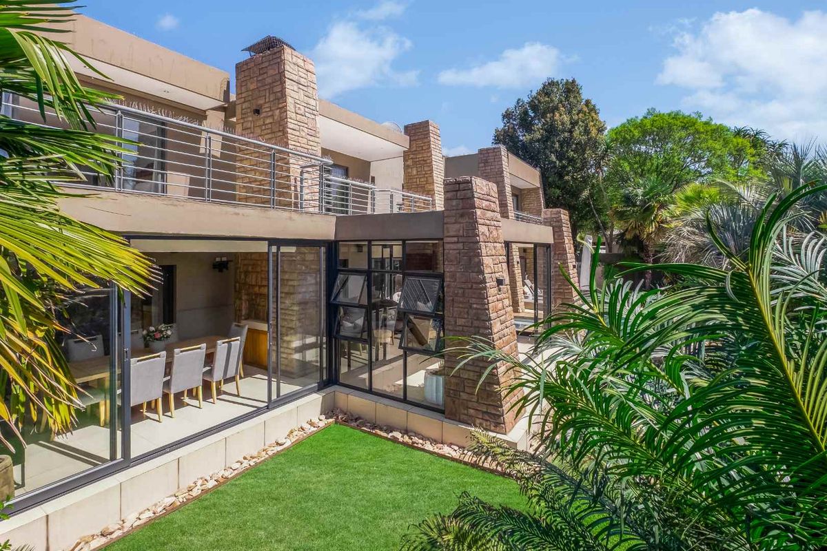 Rear view of a modern multi-level home featuring stone cladding, expansive sliding glass doors, and a spacious upper-level balcony overlooking a manicured garden.