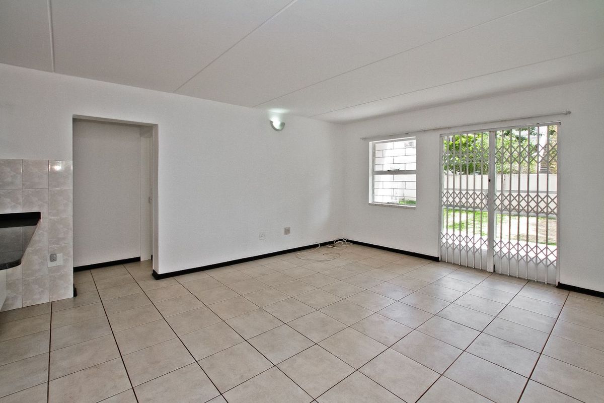A bright, empty living area featuring neutral tiled flooring, a security gate leading to the garden, and large windows with burglar bars.