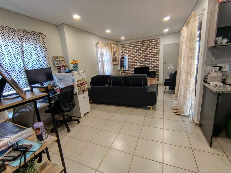 An open-plan living area featuring tiled floors, recessed lighting, and a decorative exposed brick feature wall, seamlessly connecting to the kitchen space.