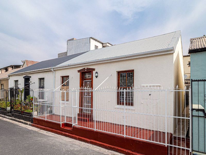 Front facade of a traditional cottage featuring a corrugated metal roof, secure metal fencing, and timber-framed windows with security bars.