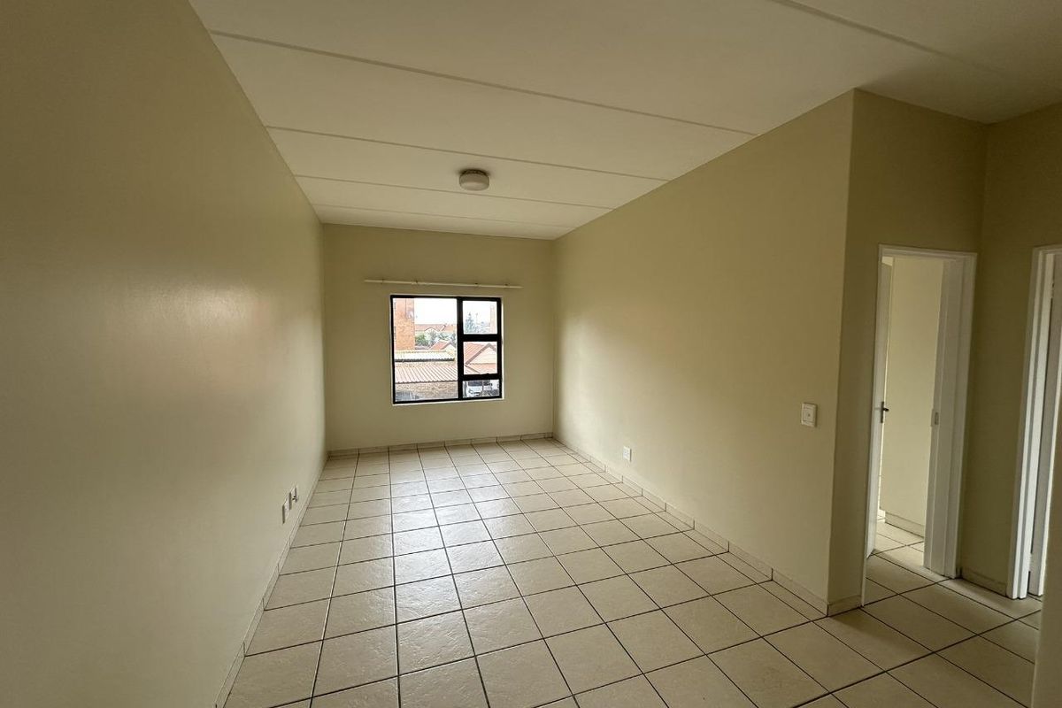 An empty, neutrally painted living area featuring tiled flooring, a large window providing natural light, and access to adjacent rooms.