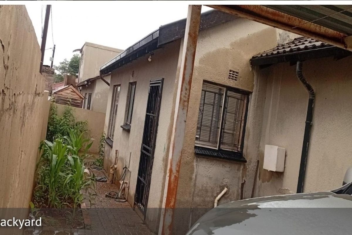 Rear exterior view of the property showing a paved walkway, security features, and signs of significant exterior wall maintenance required.