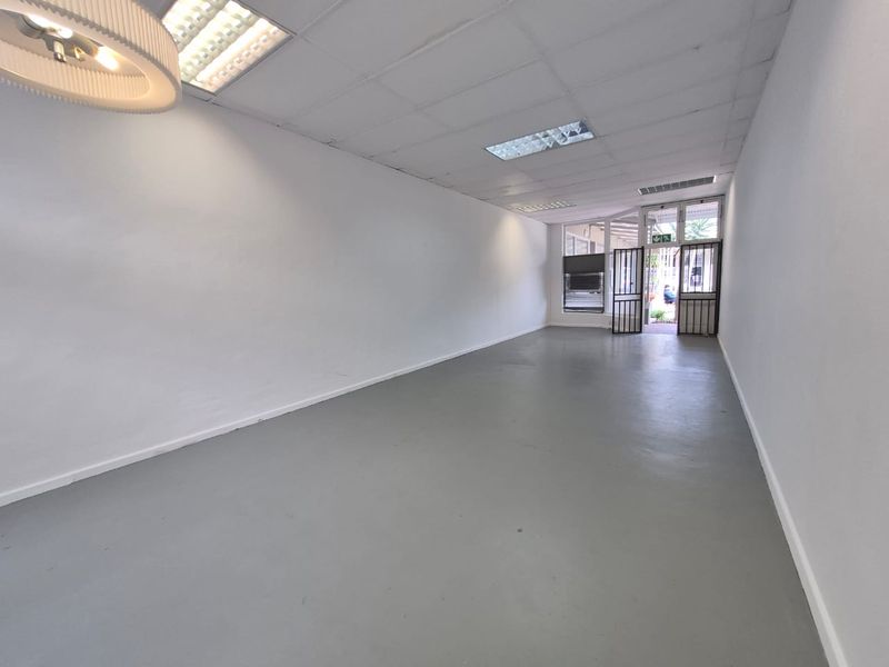 A vacant, open-plan retail space featuring a neutral painted concrete floor, suspended ceiling with recessed lighting, and a secure glass storefront entrance.