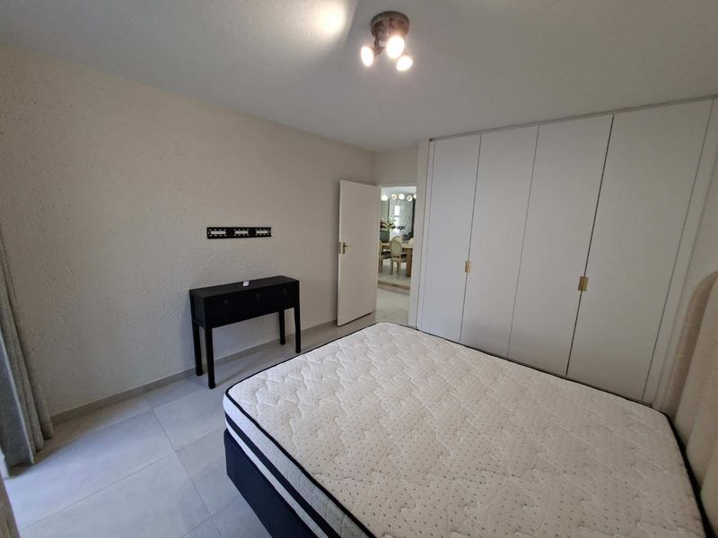 A bedroom featuring built-in cupboards, tiled flooring, and a ceiling light fixture, with a view into the adjacent dining area.