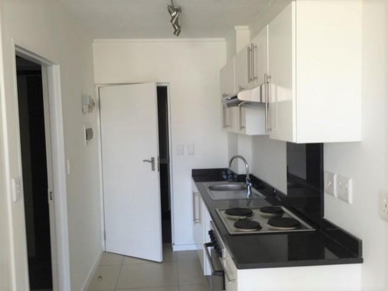 A clean, modern galley-style kitchen featuring white cabinetry, black granite countertops, a built-in electric hob, oven, and stainless steel sink.