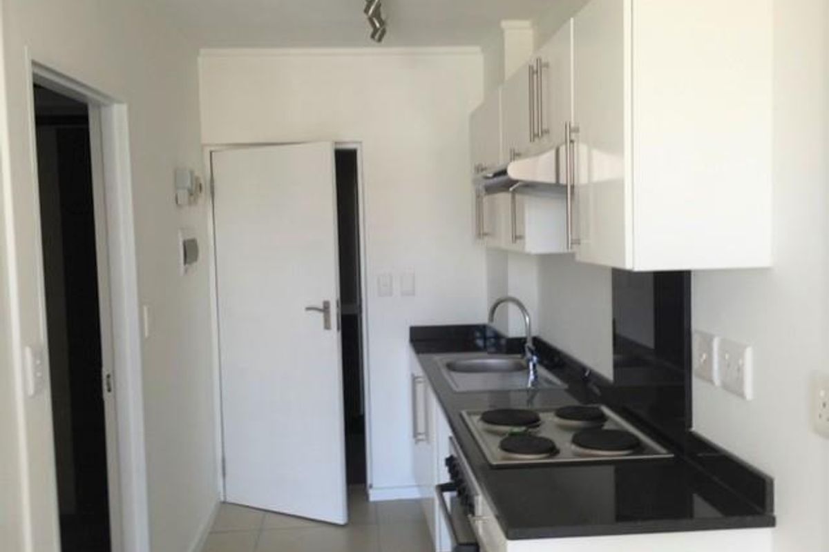 A clean, modern galley-style kitchen featuring white cabinetry, black granite countertops, a built-in electric hob, oven, and stainless steel sink.