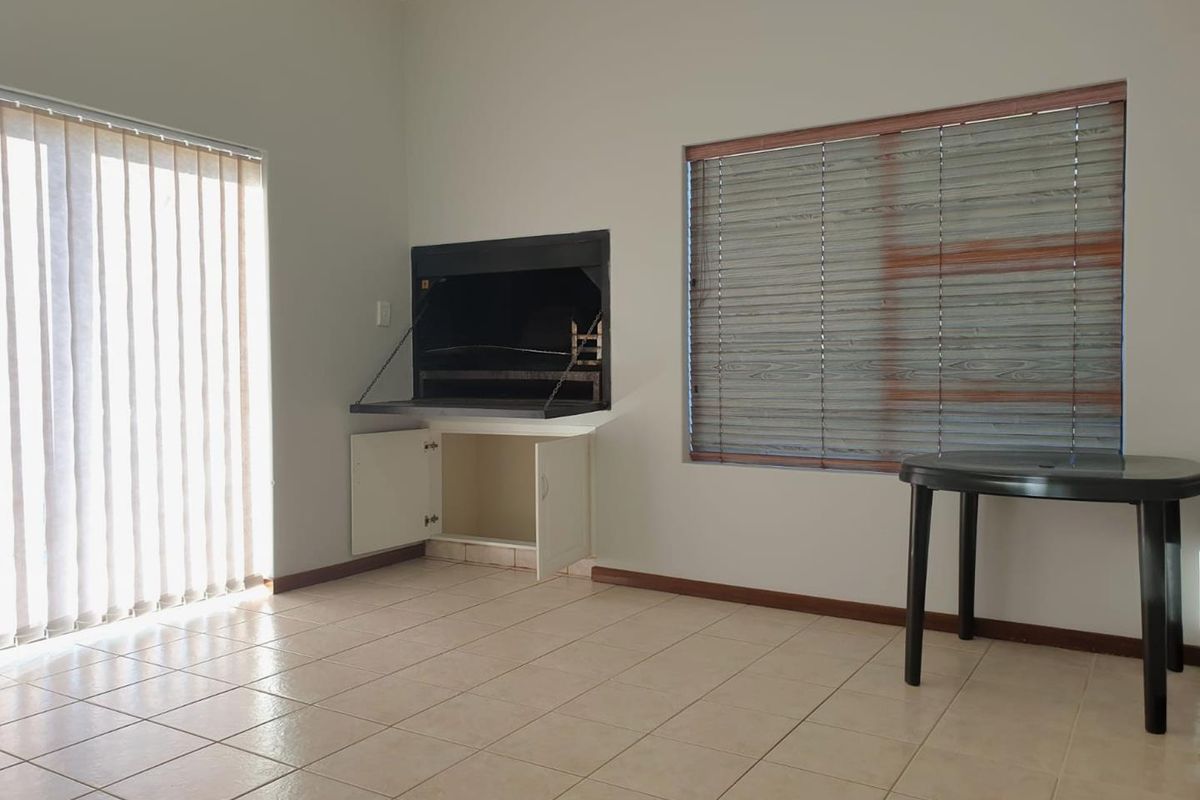 A spacious living area featuring a built-in indoor braai, tiled flooring, and large windows with blinds, offering a versatile space for entertaining.