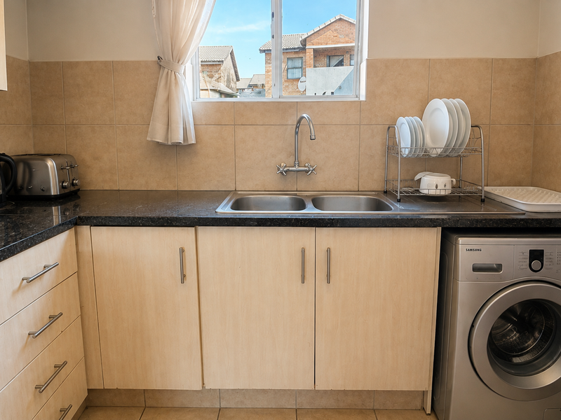 A functional kitchen featuring granite countertops, double basin sink, built-in oven, and light wood-finish cabinetry with tiled backsplash and flooring.