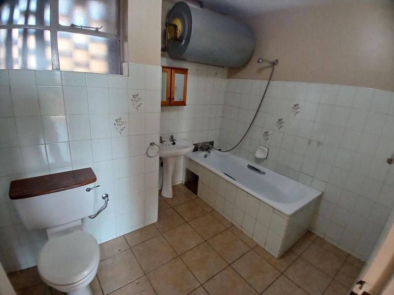 A functional bathroom featuring a pedestal basin, bathtub with shower mixer, and an exposed electric water heater, requiring cosmetic updates to modernize the space.