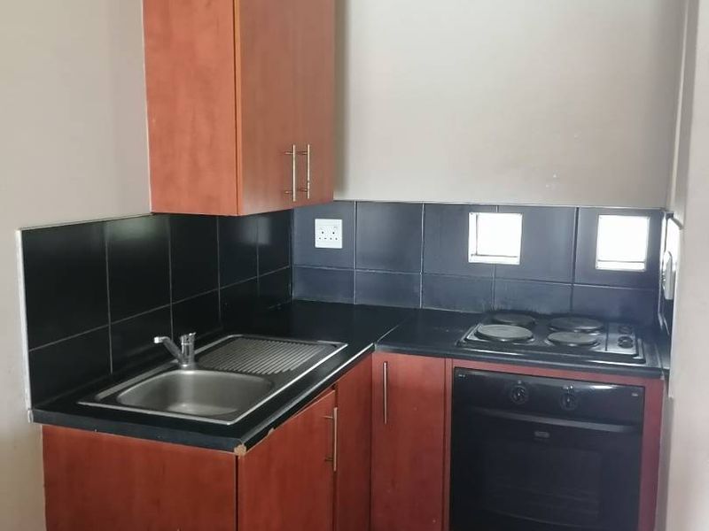 Compact kitchen featuring laminate cabinetry, a stainless steel sink, built-in oven, electric hob, and a black tiled splashback.