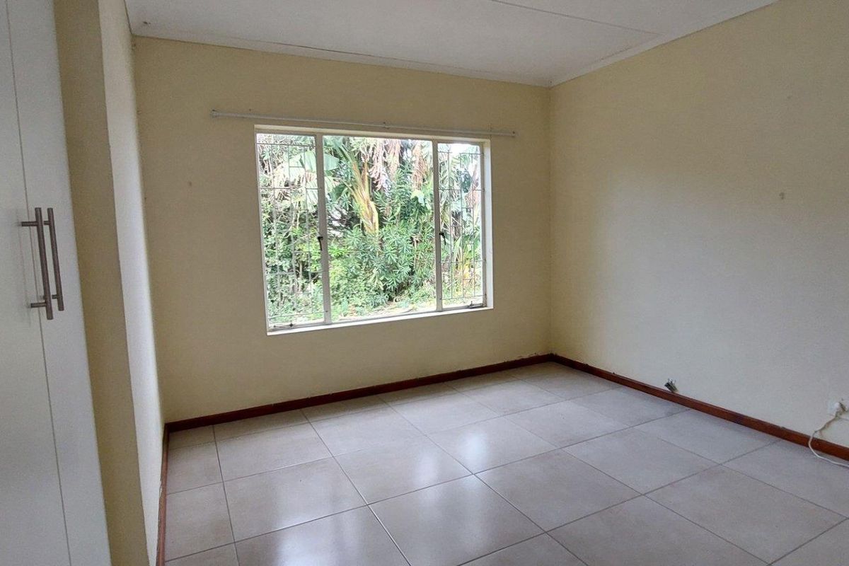 A bright, empty bedroom featuring neutral tiled flooring, built-in wardrobes, and a large window overlooking the garden.