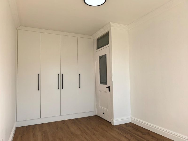 A bright, empty bedroom featuring modern white built-in wardrobes, wood-look laminate flooring, and a classic internal door with a glass panel.
