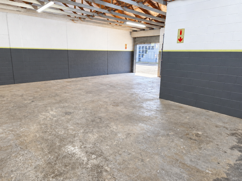 Spacious industrial garage featuring exposed timber roof trusses, durable concrete flooring, and painted block walls with a roller shutter door entrance.